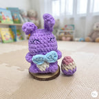 A crocheted purple bunny figurine with a light blue bow and white wing details, standing on a small wooden slice next to a matching crocheted egg, set on a beige play mat in a softly blurred room.