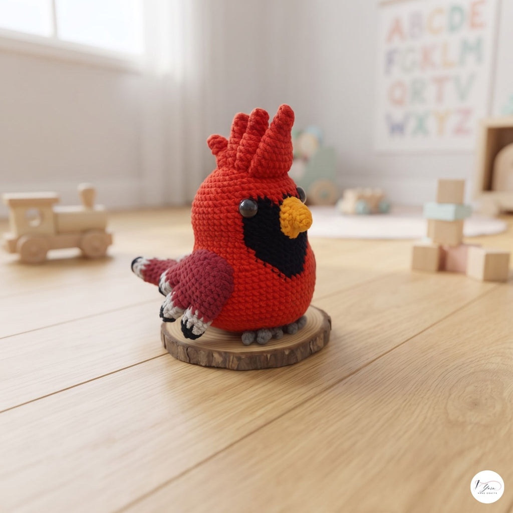 A crocheted cardinal bird figurine in bright red, with a black chest, yellow beak, and pink tail, standing on a round wooden slice base on a light wood floor. The background shows a softly blurred nursery or playroom setting with toys and a letter board.