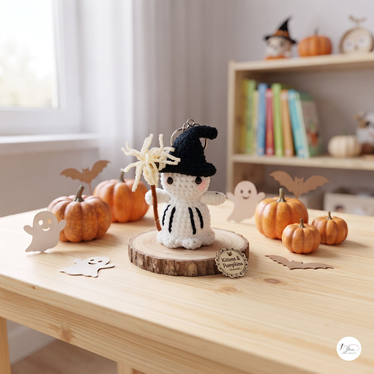 A crocheted white ghost decoration wearing a black witch hat and holding a spiderweb wand, displayed on a wooden slice stand on a light wood table. The scene is decorated with small pumpkins, ghost cutouts, and bat cutouts in a cozy, autumnal setting with a blurred bookshelf in the background.