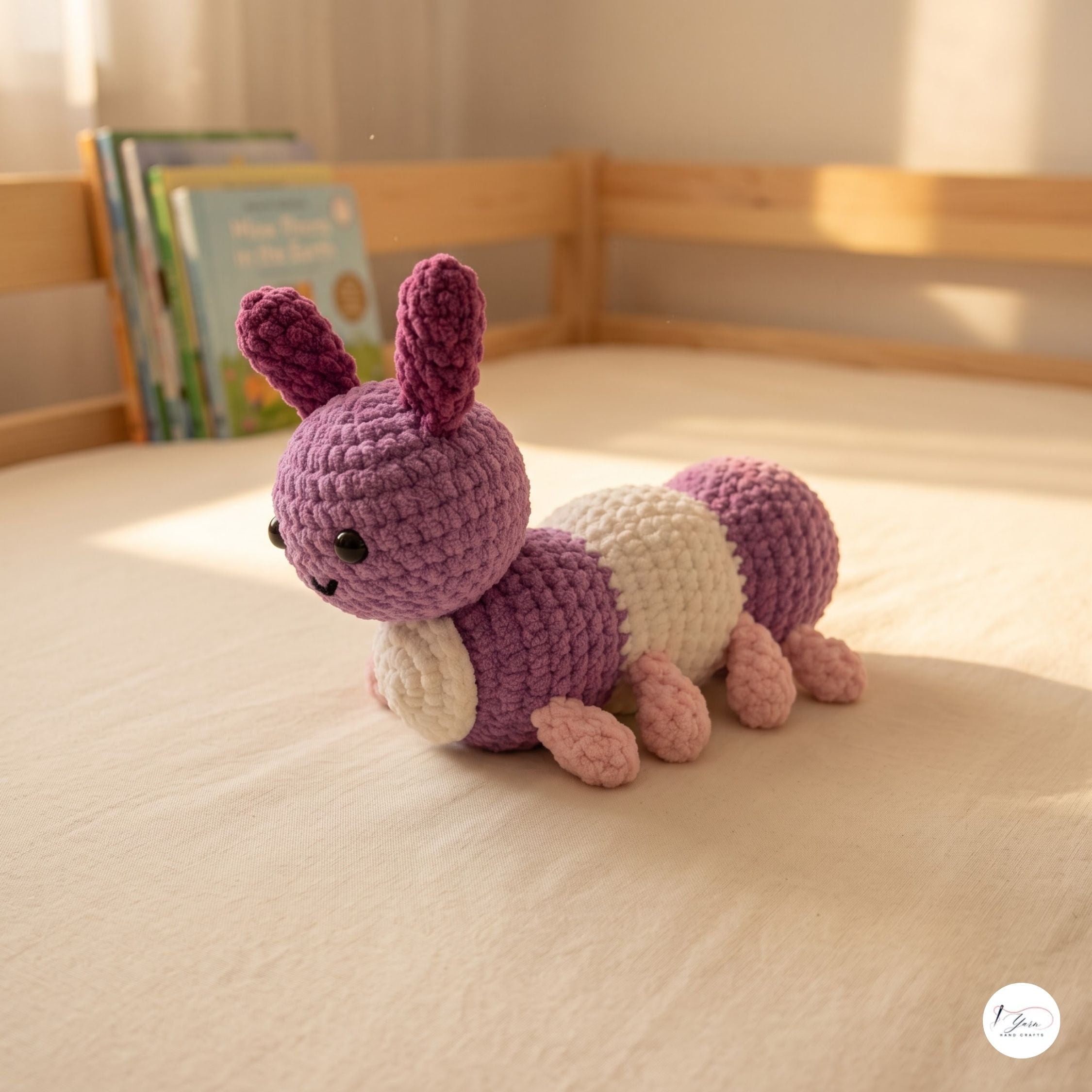 A handmade crocheted caterpillar toy in shades of purple, cream, and pink, resting on a beige bedsheet in a child's bedroom with a wooden bed frame and books in the background.