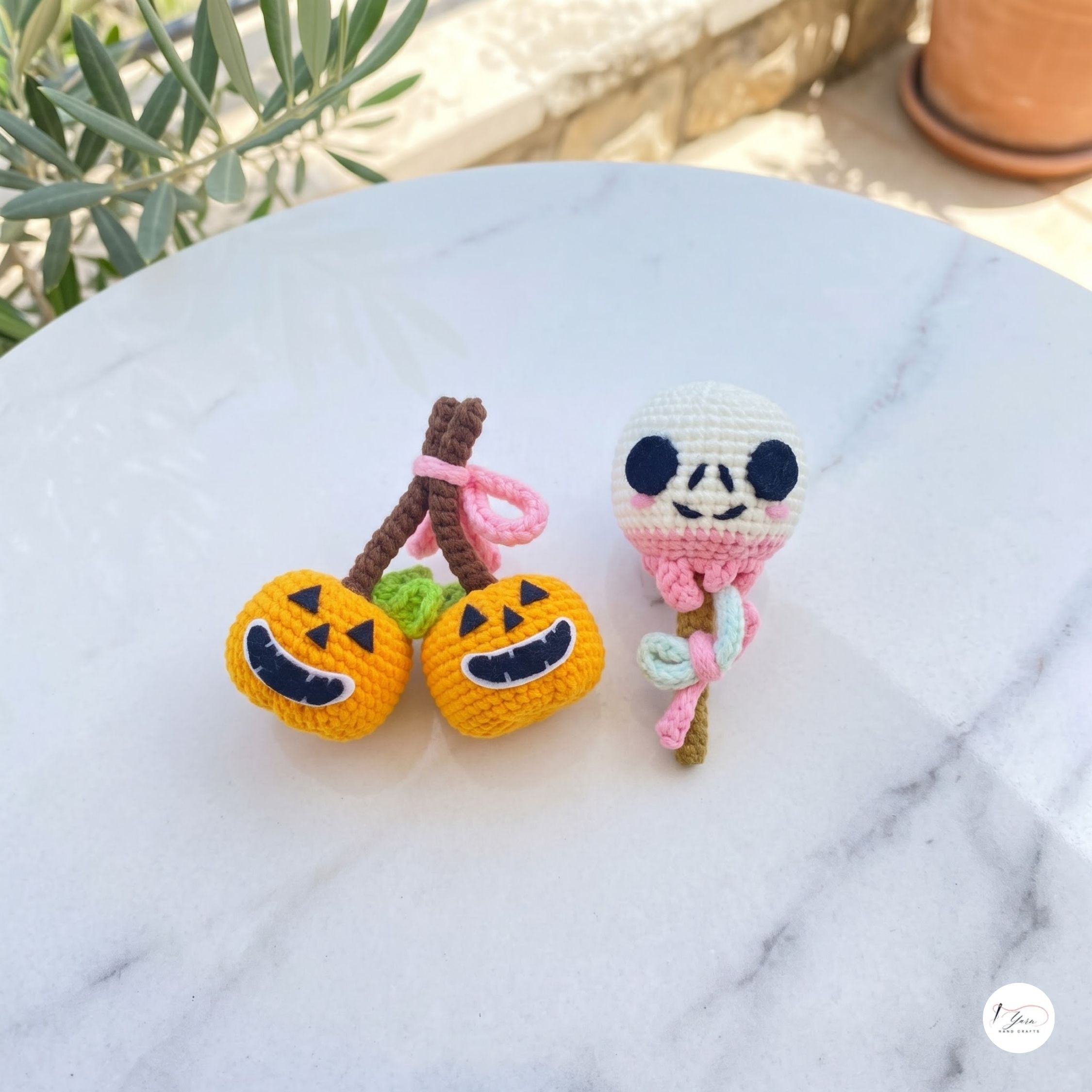 Two handmade crocheted Halloween-themed charms resting on a white marble surface outdoors. One charm features two orange jack-o'-lantern pumpkins with brown stems and pink ties, and the other is a white ghost with black embroidered eyes and mouth, pink details, and a brown stem.