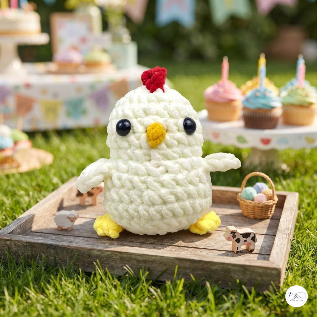 A crocheted white chick toy with a red comb, yellow beak and feet, sitting on a wooden tray outdoors on green grass. Cupcakes and small animal figurines are blurred in the background.