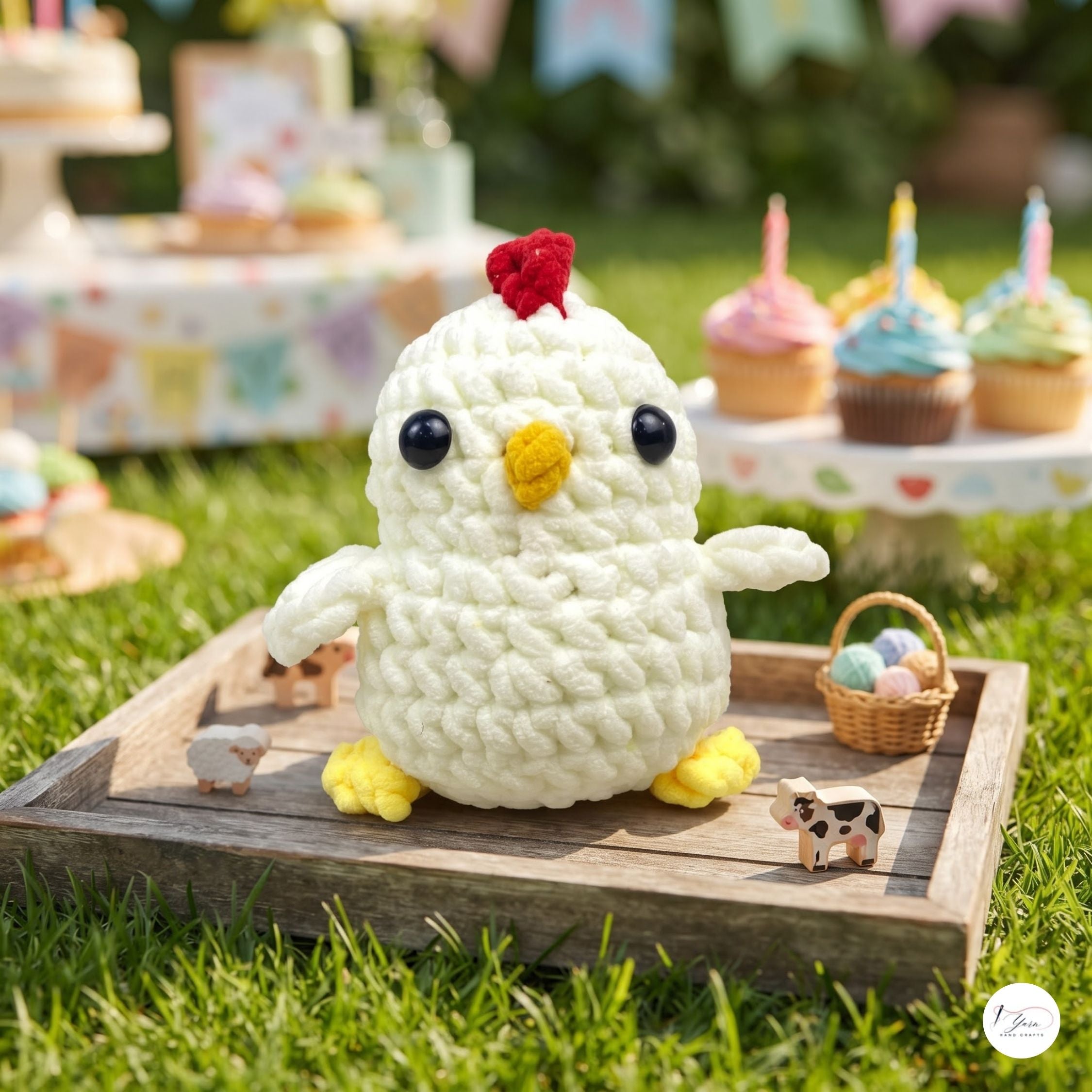 A crocheted white chick toy with a red comb, yellow beak and feet, sitting on a wooden tray outdoors on green grass. Cupcakes and small animal figurines are blurred in the background.