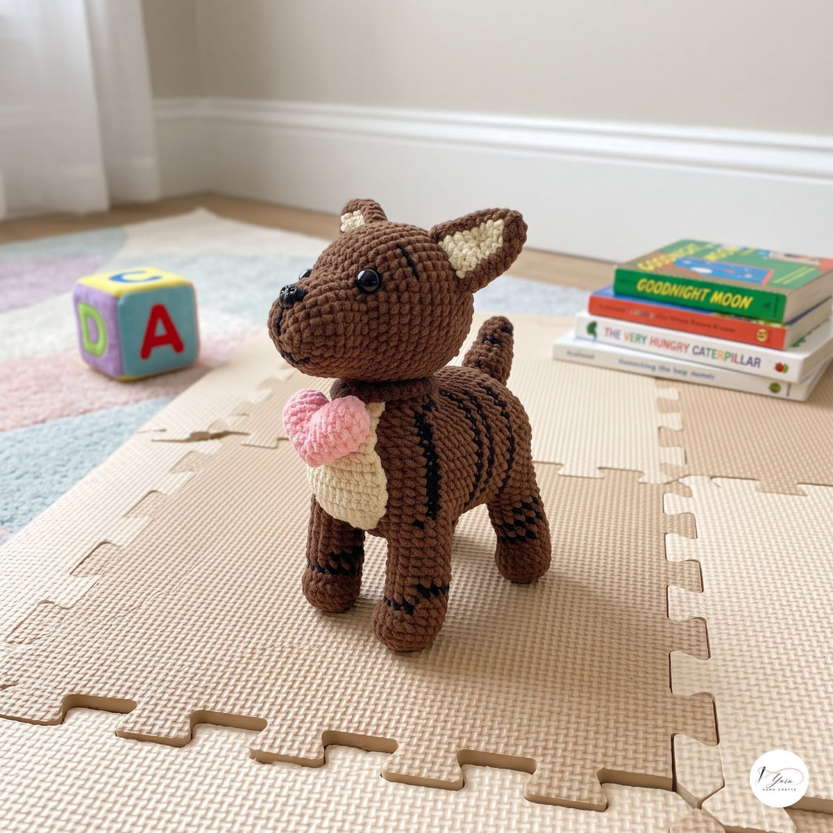A crocheted plush toy dog standing on beige interlocking foam play mats in a bright room. The toy is brown with black stripes, has cream inner ears, and holds a pink heart-shaped accessory.