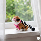 A crocheted cat toy sits on a white windowsill with sheer curtains and a blurred green garden background. The toy is orange with black stripes, white paws and face, and wears a pink collar.