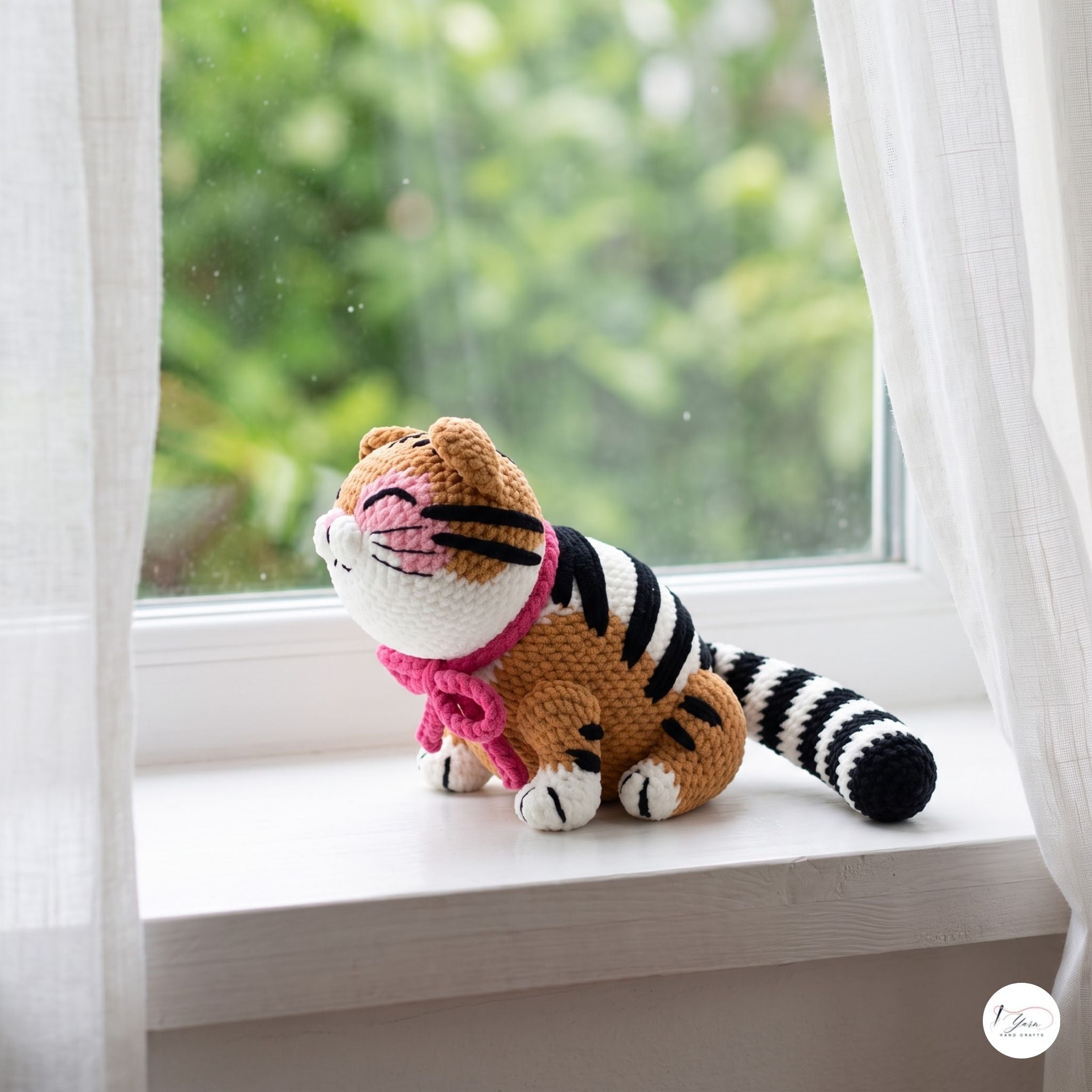 A crocheted cat toy sits on a white windowsill with sheer curtains and a blurred green garden background. The toy is orange with black stripes, white paws and face, and wears a pink collar.