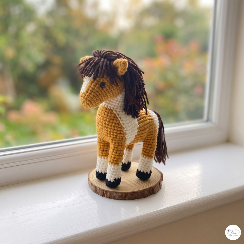 A crocheted toy horse with a brown and cream body, dark brown yarn mane and tail, standing on a round wooden slice base on a white windowsill with a blurred garden view in the background.