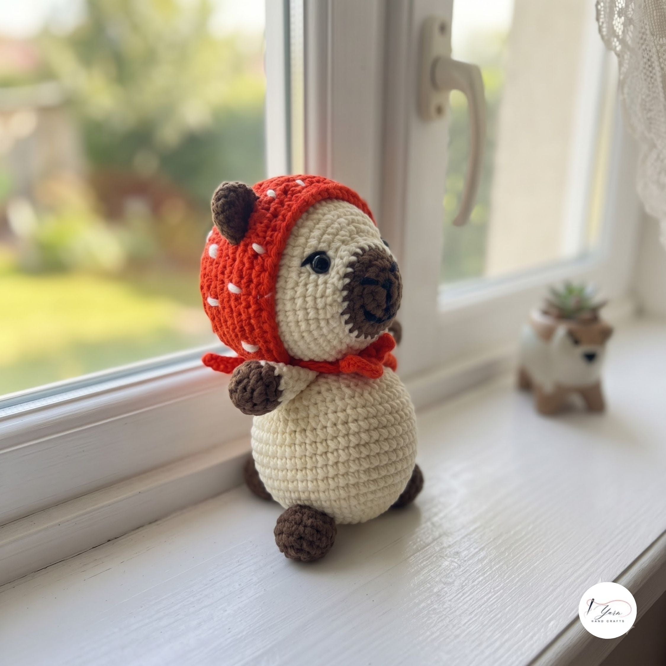 A crocheted pig doll wearing a red hat with white spots and a red scarf, sitting on a white windowsill with a blurred outdoor garden background. A small plant pot with a pig figurine is visible in the background.