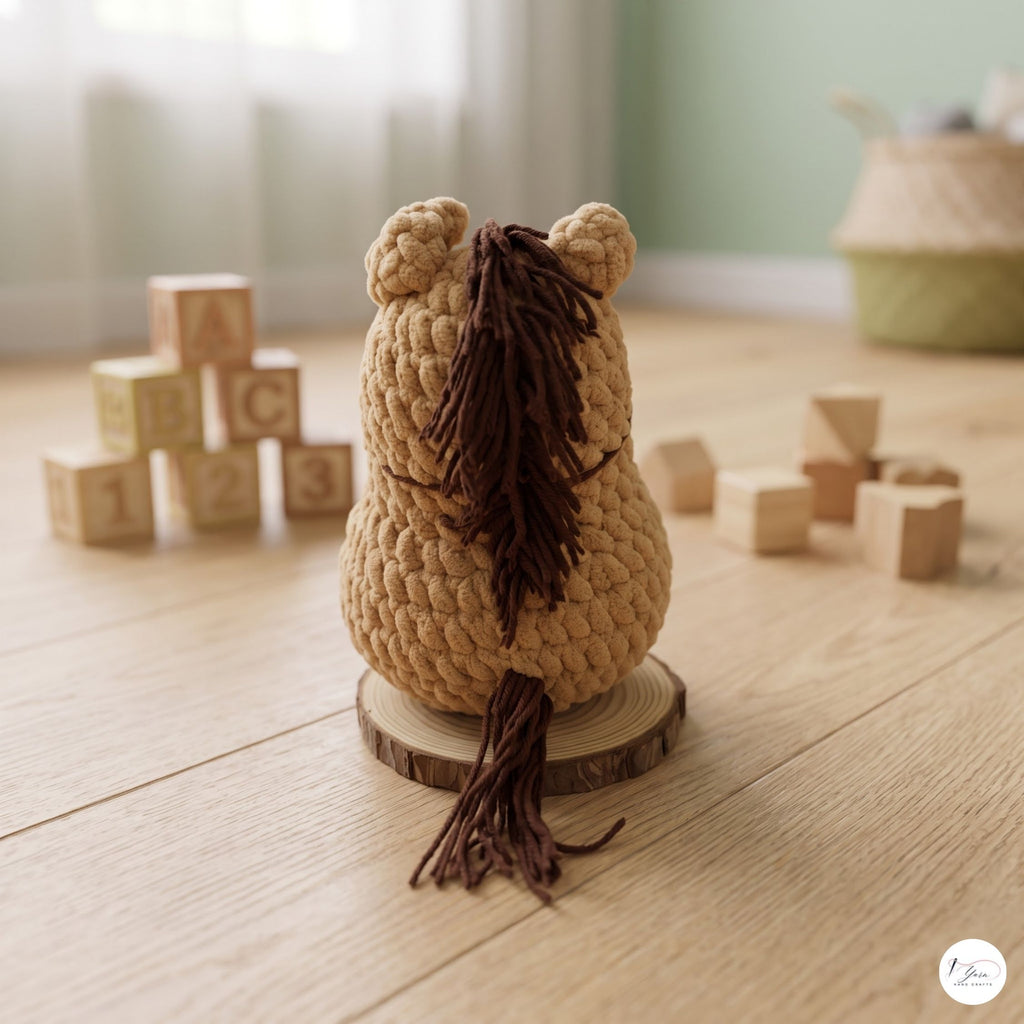 A handmade crocheted plush horse toy, shown from the back, sitting on a round wooden slice coaster on a light wood floor. The toy is made of beige yarn with a dark brown mane and tail.
