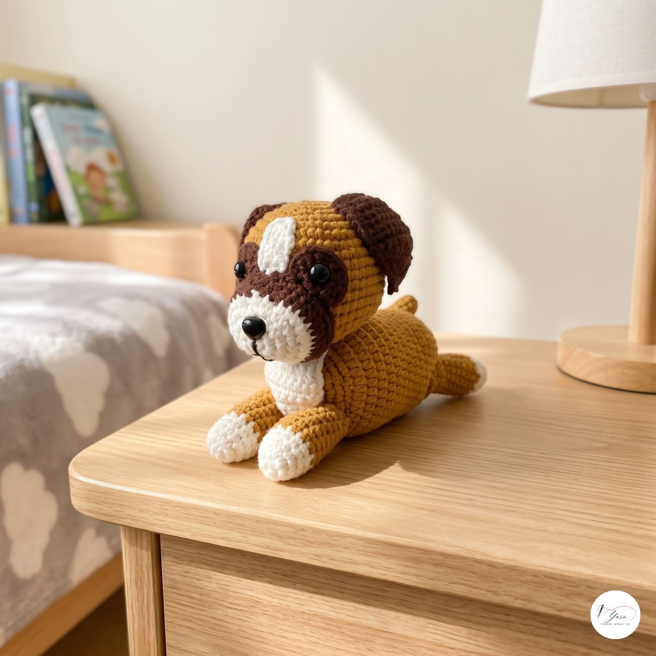 A crocheted plush puppy toy in brown, white, and golden yellow sits on a light wood bedside table in a softly lit bedroom. A lamp is visible on the right, and a bed with a gray cloud-patterned blanket is in the background.