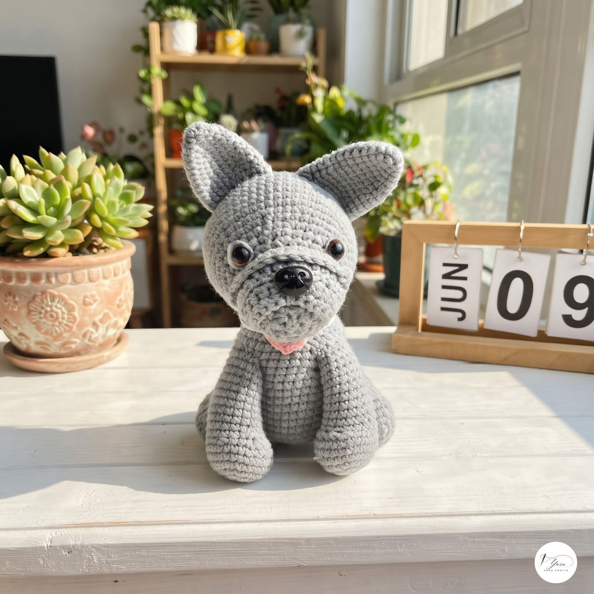 A crocheted gray dog plush toy with large upright ears, black eyes, a black nose, and a small pink collar, sitting on a white tabletop in a sunlit room with plants and a wooden calendar in the background.