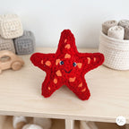 A red crocheted starfish toy with a smiling face and blue eyes sits on a light wood shelf in a nursery-like setting, surrounded by soft toys and storage baskets.