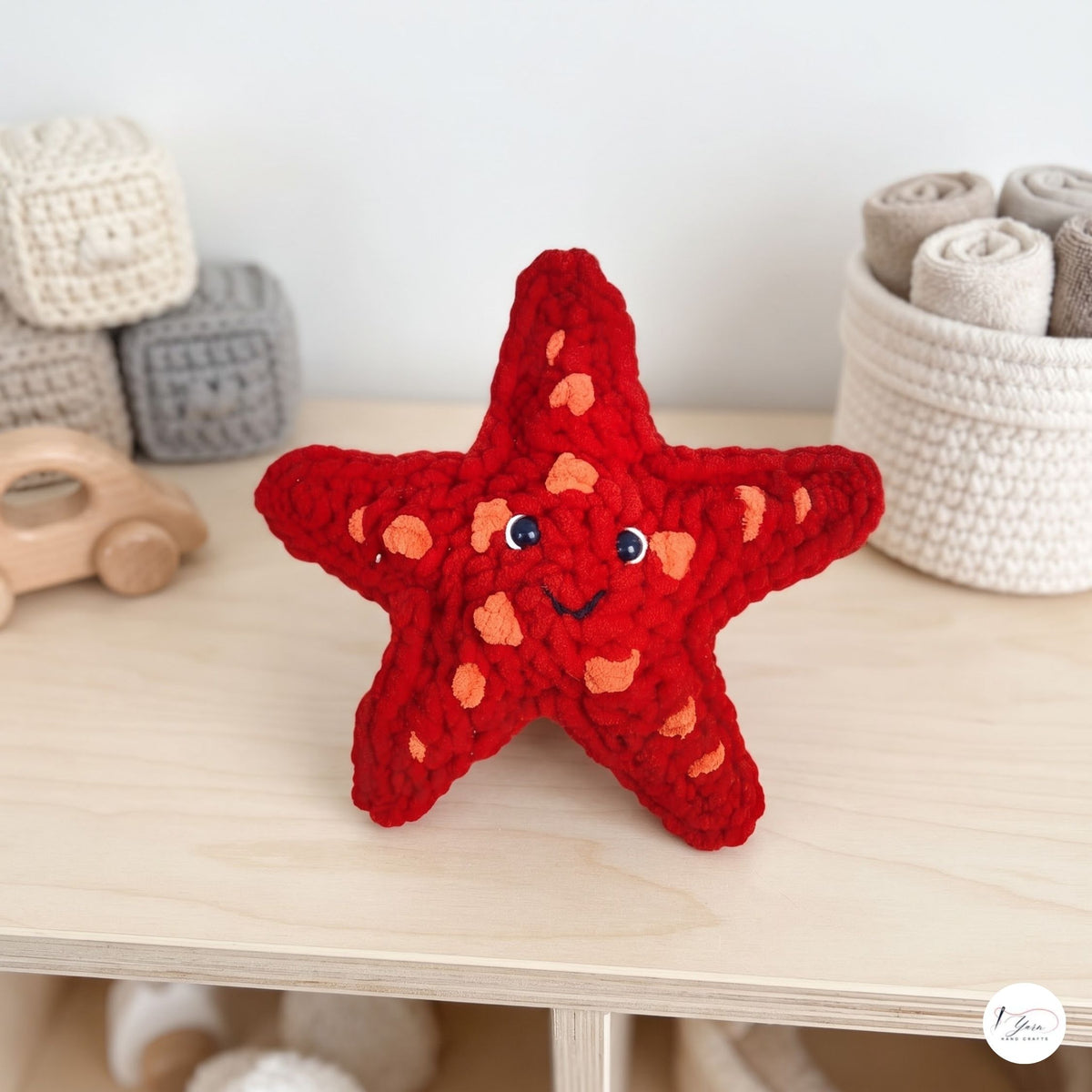 A red crocheted starfish toy with a smiling face and blue eyes sits on a light wood shelf in a nursery-like setting, surrounded by soft toys and storage baskets.