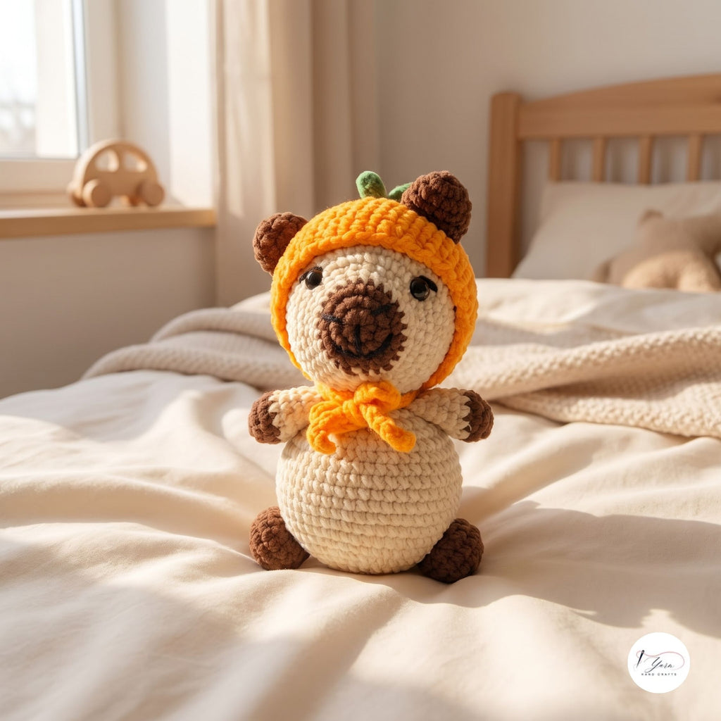 A crocheted plush toy bear wearing an orange pumpkin hat and bow tie, sitting on a cream-colored bed in a softly lit bedroom.