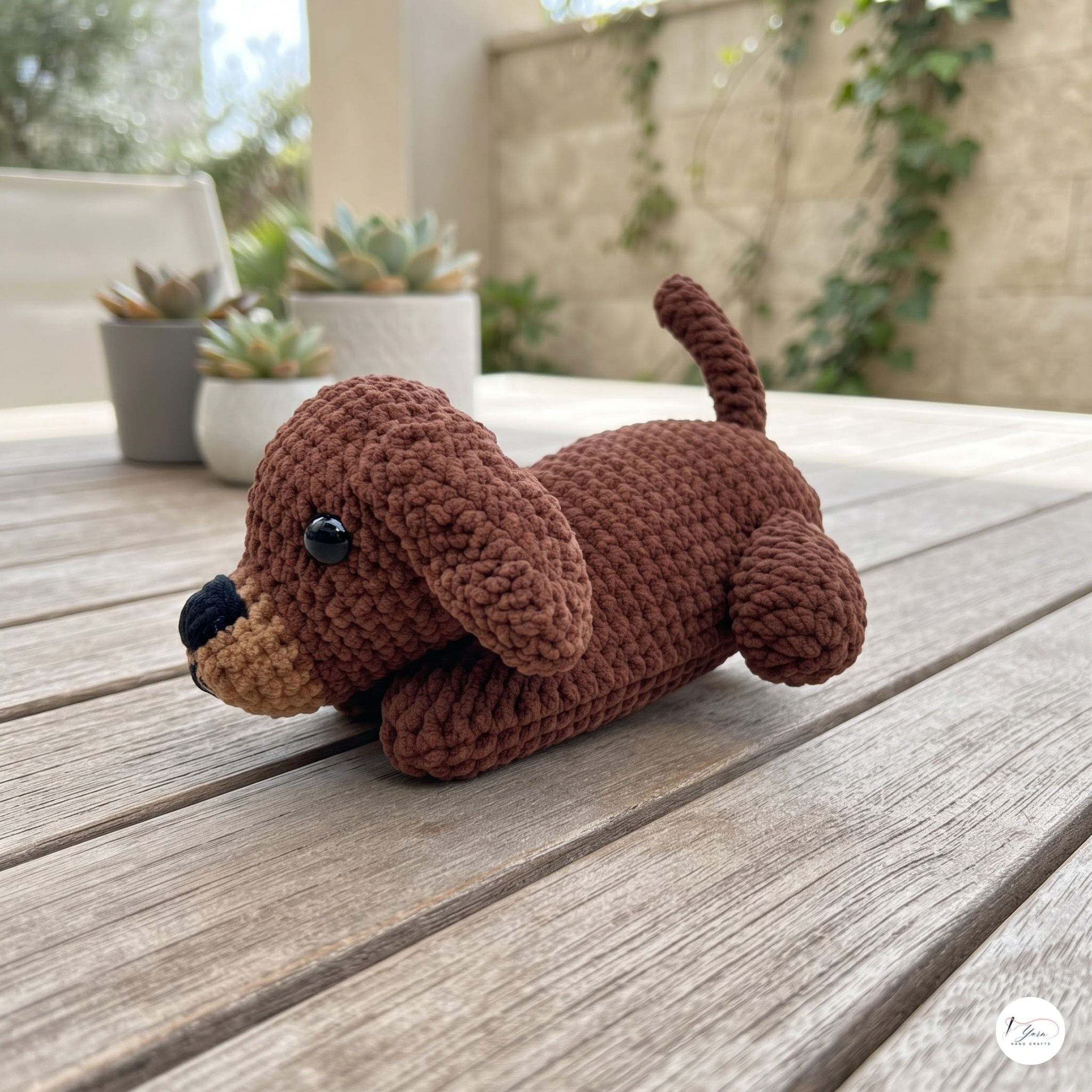 A brown crocheted plush puppy toy with a black nose and glossy black eye, lying on a weathered wooden deck table with potted succulents in the background.