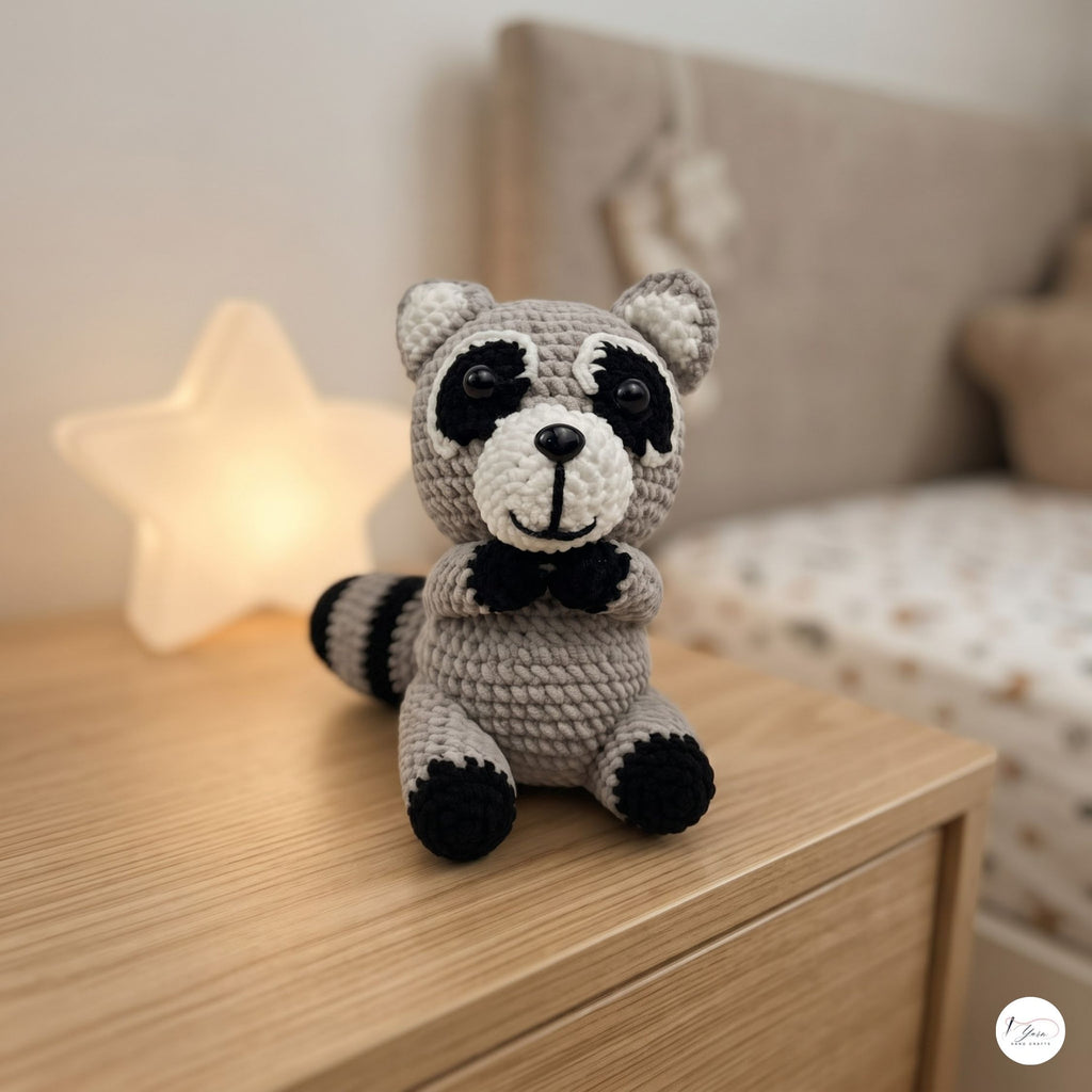 A crocheted raccoon plush toy sits on a light wood bedside table in a softly lit bedroom. The toy is gray and black with embroidered facial details and a small bow tie.