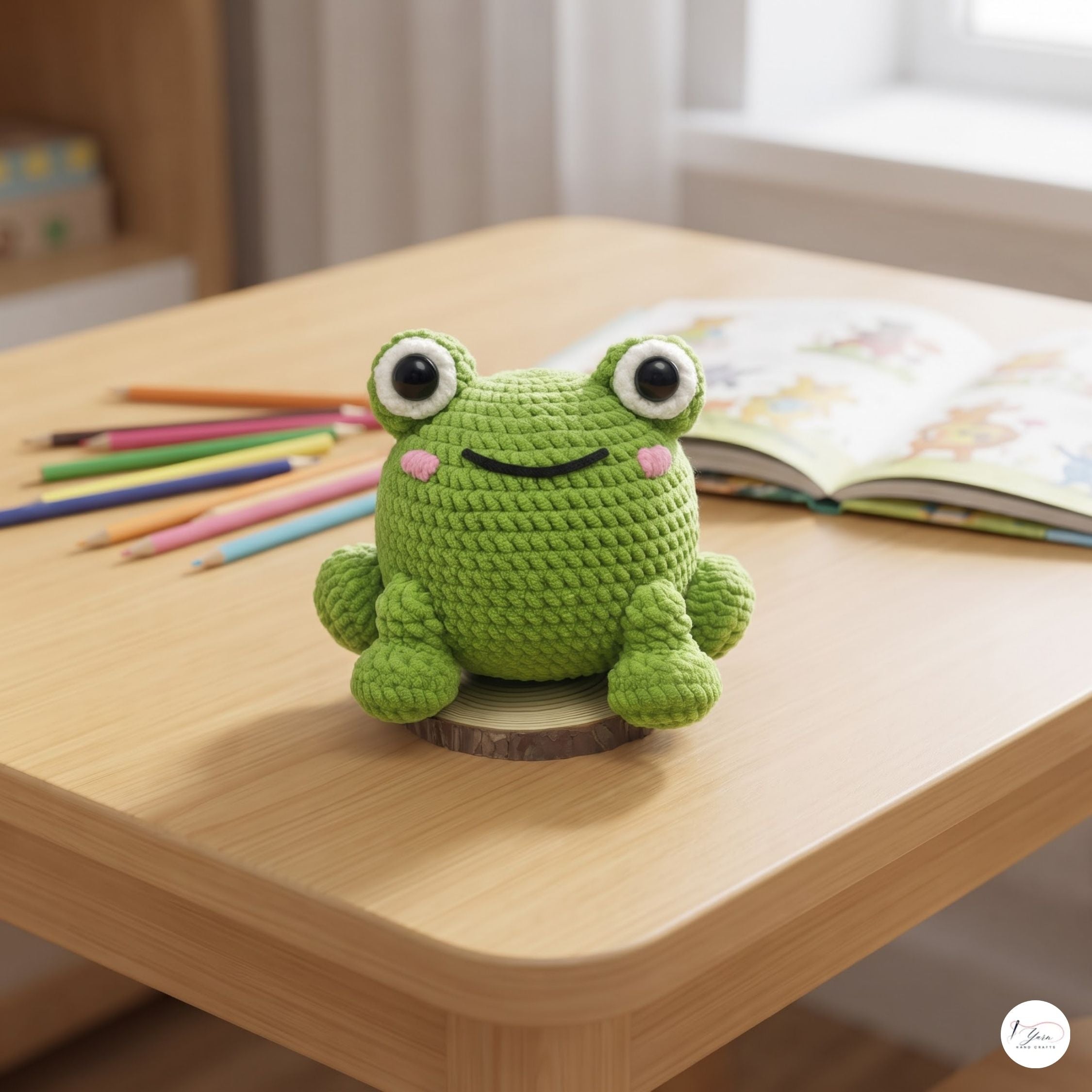 A green crocheted frog toy sitting on a light wood table, with colored pencils and an open book in the background.