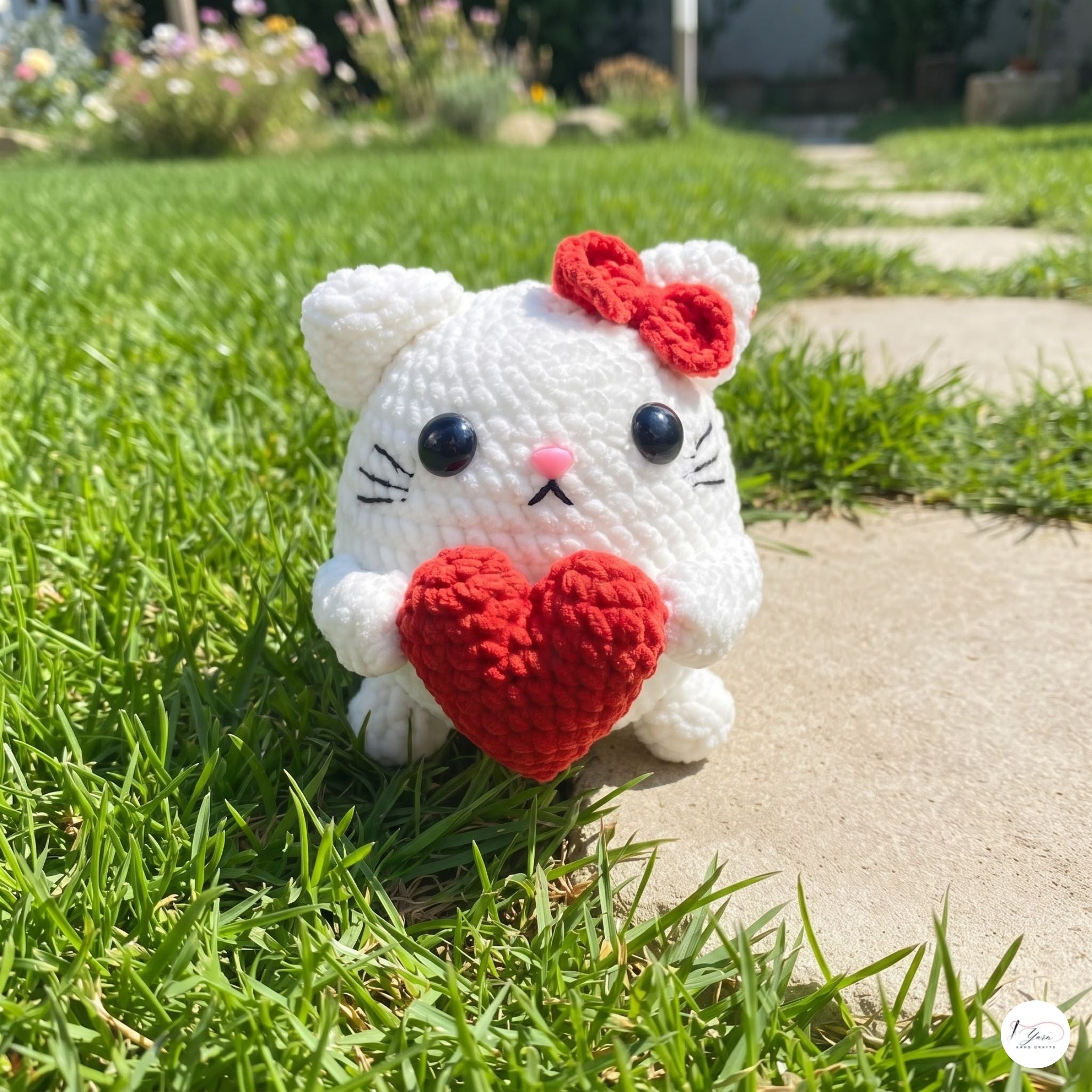 A white crocheted cat amigurumi toy holding a red heart, sitting on a green grass lawn next to a concrete path in a backyard.