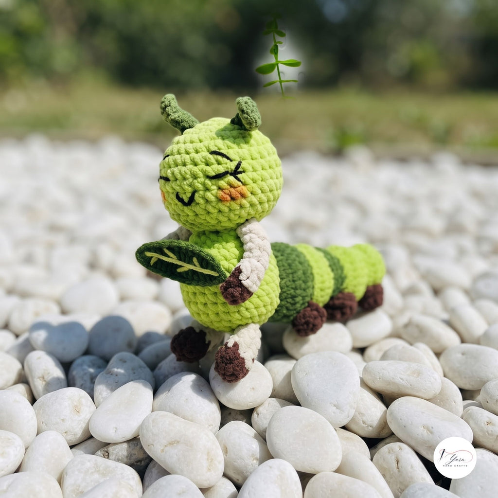 A crocheted caterpillar toy standing on white pebbles outdoors, with a blurred green garden background. The toy is lime green with darker green stripes, cream-colored legs, and small brown feet.