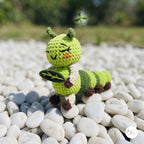 A crocheted caterpillar toy standing on white pebbles outdoors, with a blurred green garden background. The toy is lime green with darker green stripes, cream-colored legs, and small brown feet.