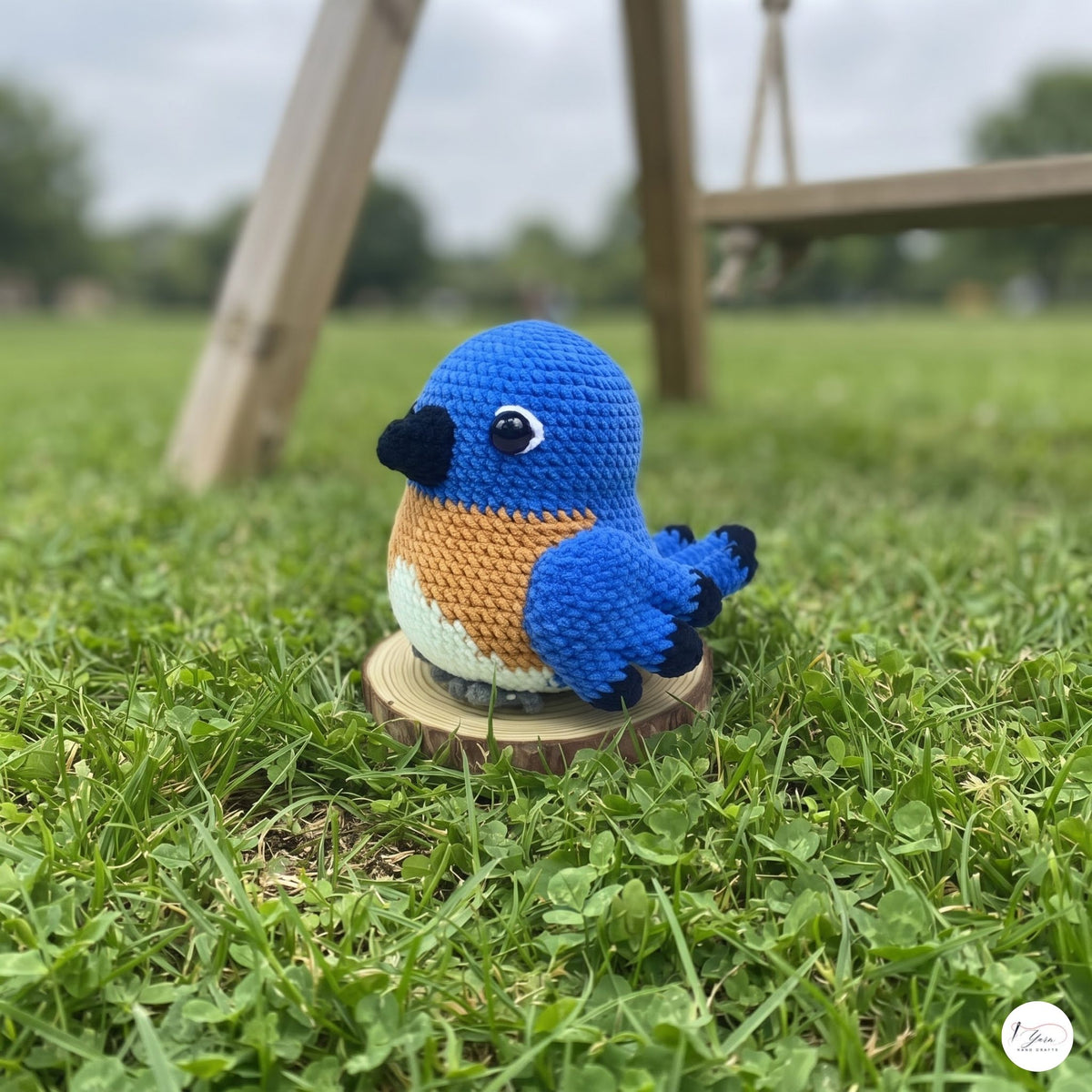 A crocheted bird toy in bright blue, orange, and white sits on a small wooden slice stand on a grassy lawn with a blurred outdoor background.