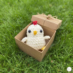 A small crocheted white chicken plush toy with a red comb and yellow beak sits inside an open kraft paper gift box on green grass.
