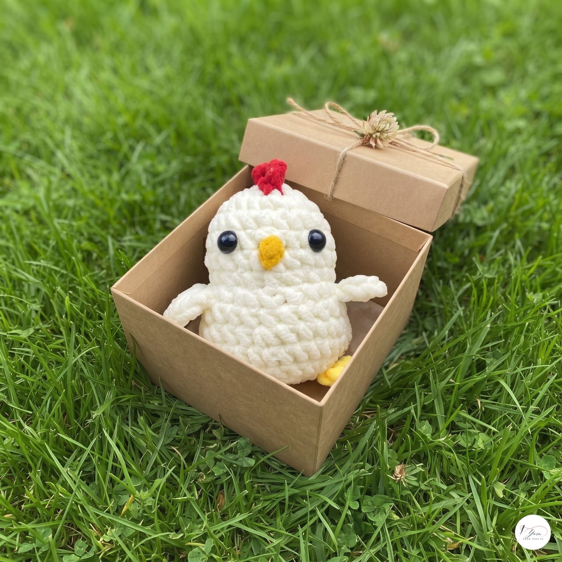 A small crocheted white chicken plush toy with a red comb and yellow beak sits inside an open kraft paper gift box on green grass.