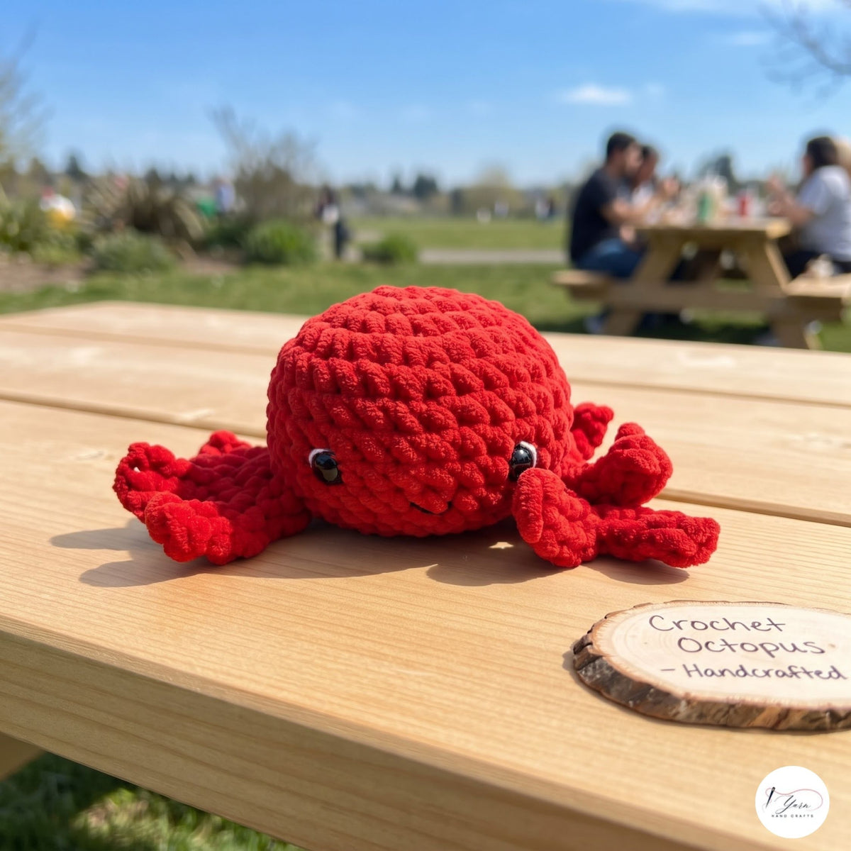 A red crocheted octopus toy sits on a light wood picnic table outdoors, with a blurred park scene and people in the background. A small sign on the table reads 'Crochet Octopus - Handcrafted' and a circular logo in the corner appears to be a maker mark.
