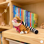 A crocheted tiger plush toy with orange and black stripes, white paws and face, and a pink bow, sitting on a wooden shelf in front of colorful children's books.