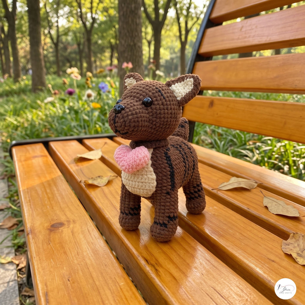 A crocheted brown bear toy holding a pink and cream ice cream cone, standing on a wooden park bench with trees in the background.
