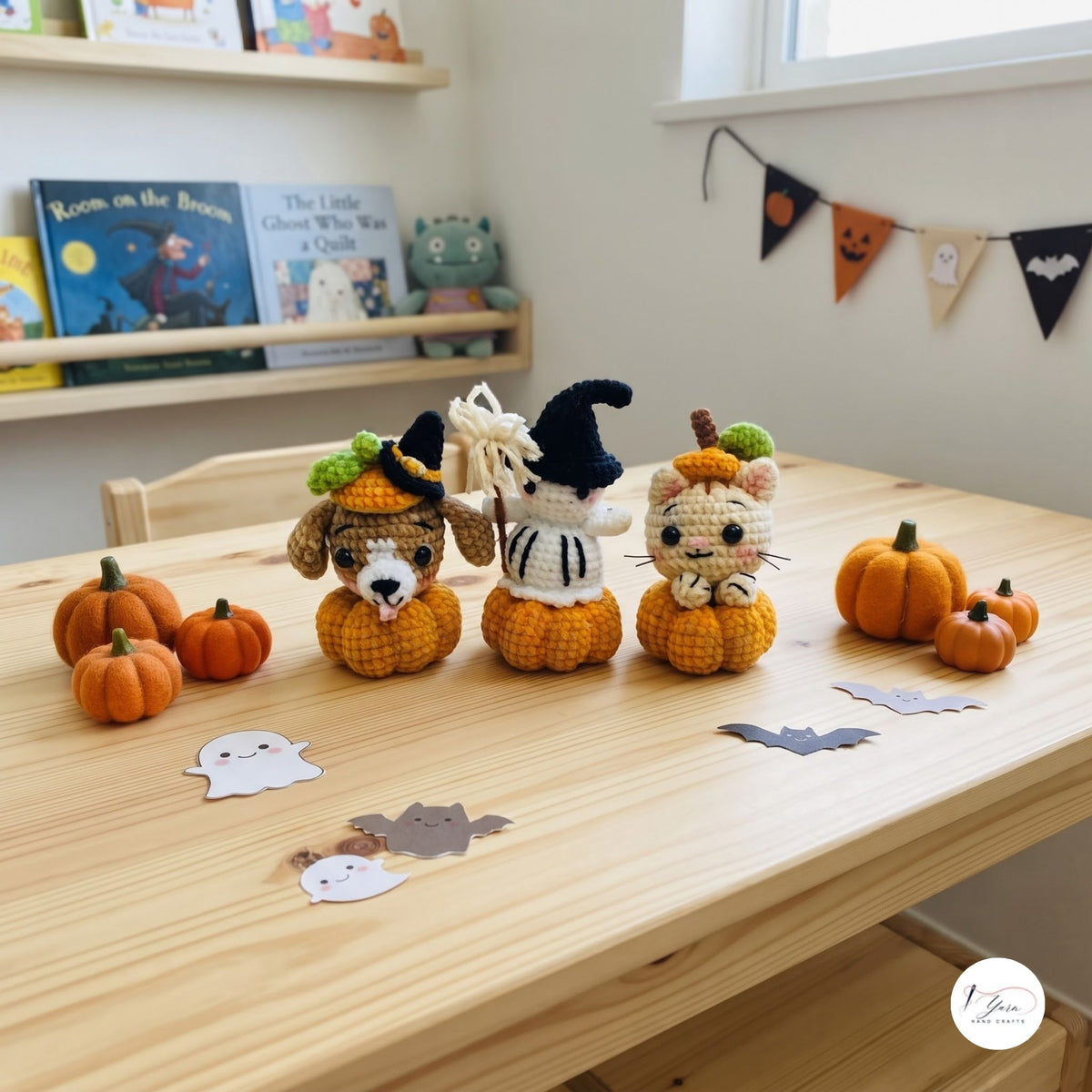 Three crocheted Halloween-themed plush toys sitting on orange yarn pumpkins on a light wood table, with small paper ghosts and bats, books on shelves, and a window in the background.