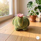 A handmade crocheted cactus plush toy in olive green with yellow spines and a pink flower on top, sitting on a wooden windowsill in a sunlit room with potted plants.