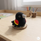 A crocheted bird figurine in black with red, yellow, and gray accents, standing on a round wooden coaster on a light wood desk.