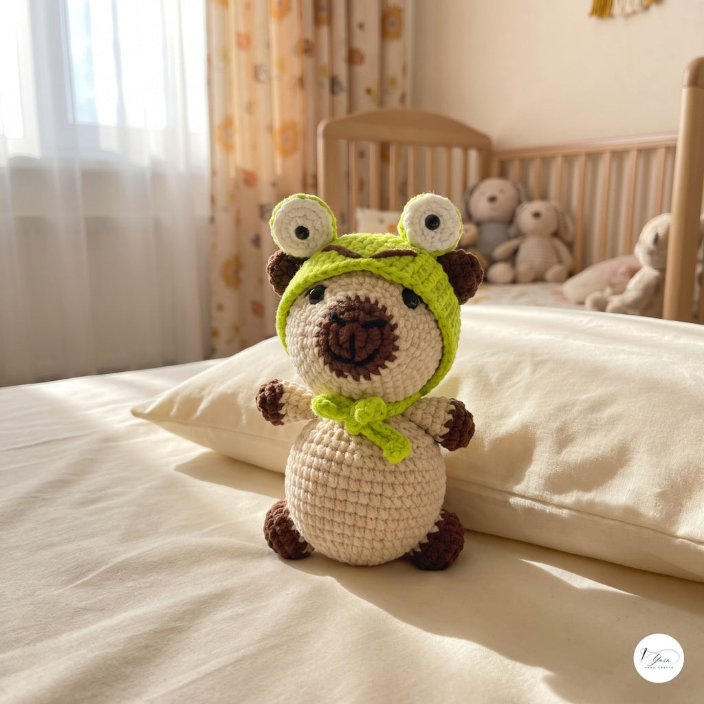A crocheted plush toy dog wearing a green frog hat and matching green scarf, sitting on a bed in a softly lit nursery. The background shows a wooden crib, patterned curtains, and stuffed toys.