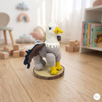 A crocheted eagle figurine with a white head, yellow beak and feet, gray body, brown and black wings, displayed on a round wood slice stand on a light wooden floor in a softly blurred children's room.