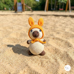 A small crocheted kangaroo toy stands in sandy ground outdoors, wearing an orange hood with long ears and a matching orange scarf. The background shows a blurred playground with trees and wooden structures.