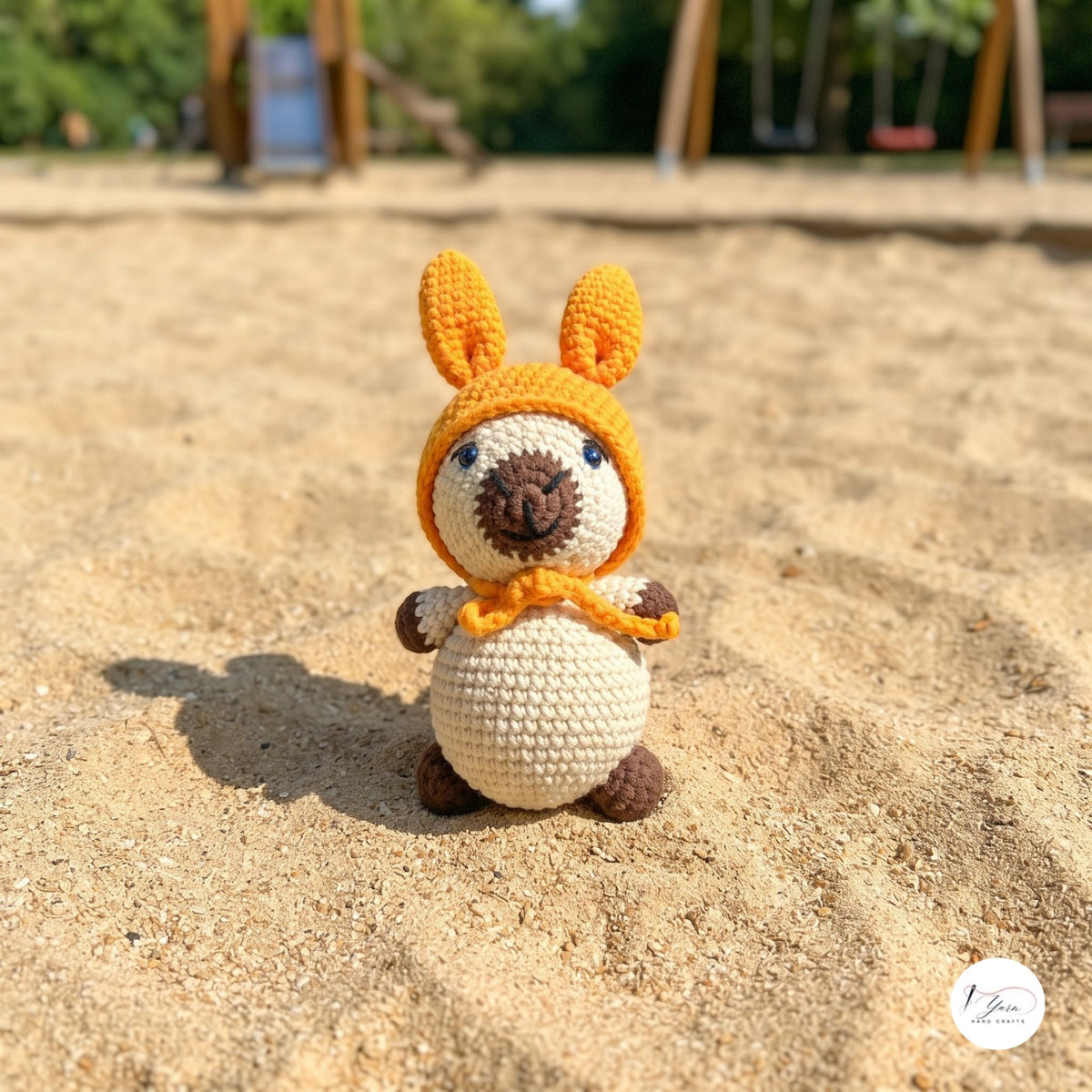 A small crocheted kangaroo toy stands in sandy ground outdoors, wearing an orange hood with long ears and a matching orange scarf. The background shows a blurred playground with trees and wooden structures.