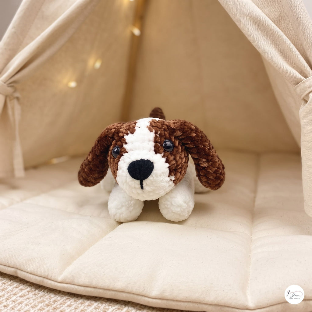 A small crocheted dog plush toy with brown and white fur sits on a cream-colored padded cushion inside a soft fabric dog teepee with warm string lights.