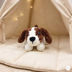A small crocheted dog plush toy with brown and white fur sits on a cream-colored padded cushion inside a soft fabric dog teepee with warm string lights.
