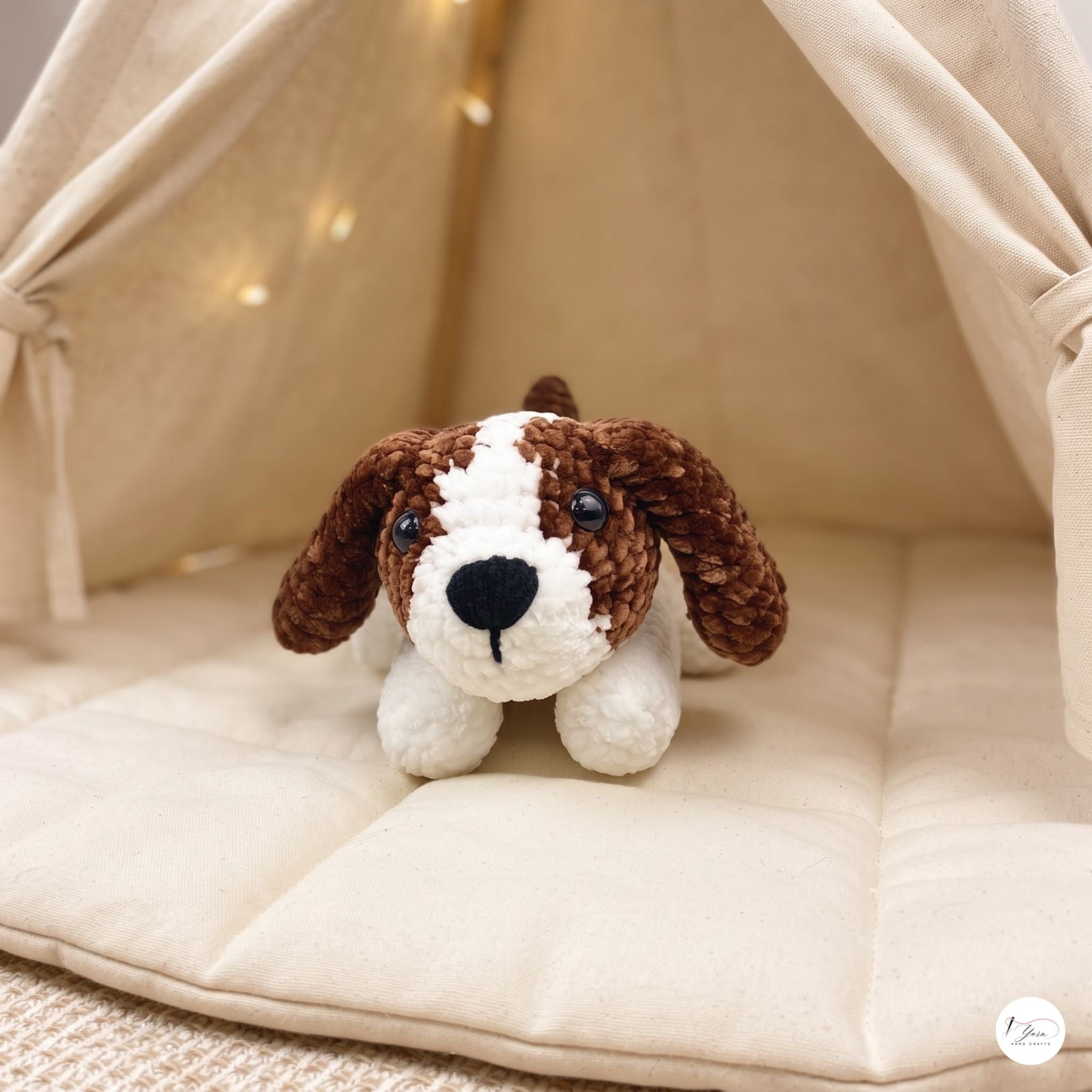 A small crocheted dog plush toy with brown and white fur sits on a cream-colored padded cushion inside a soft fabric dog teepee with warm string lights.