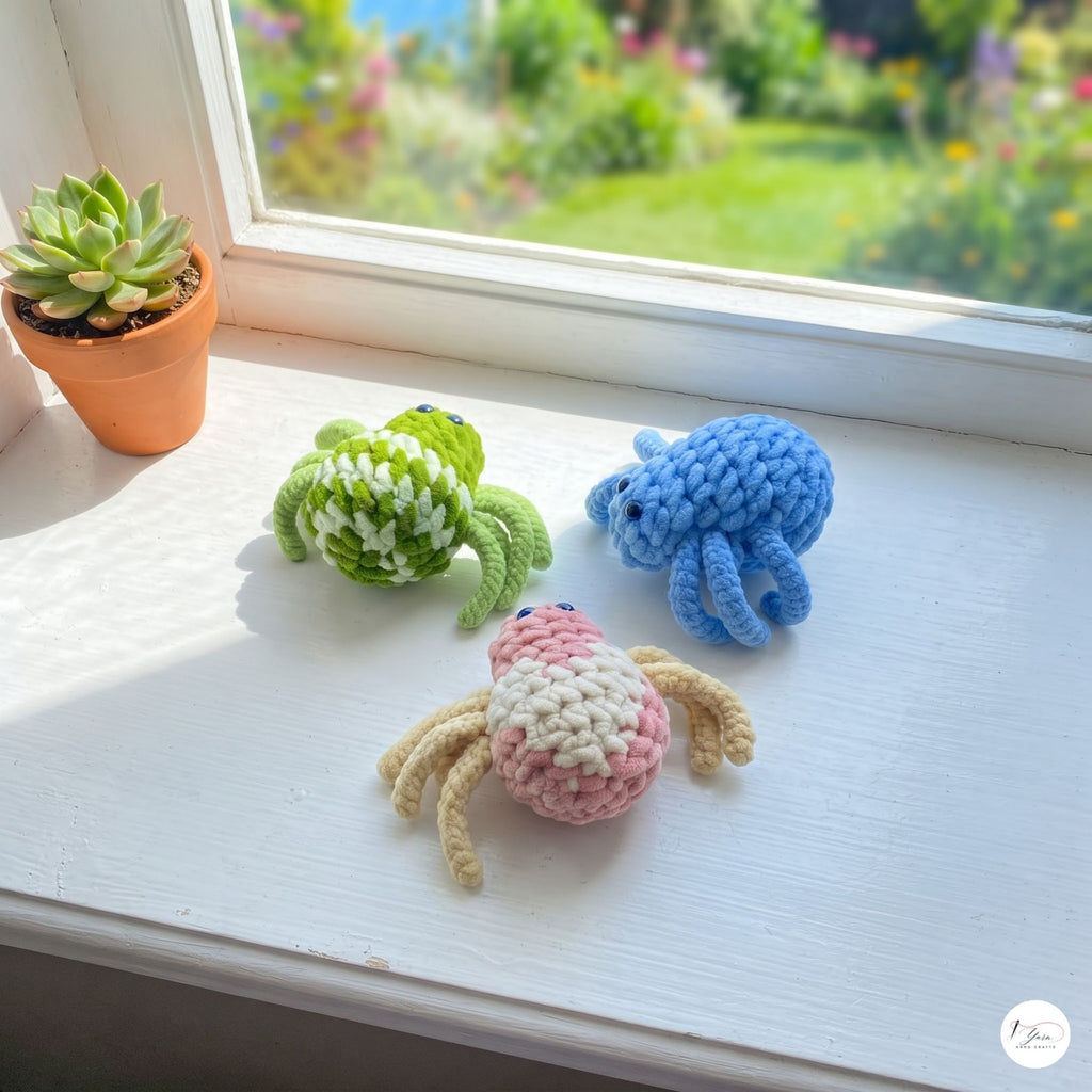 A colorful trio of plush handmade crochet spider toys in mottled green and cream, light blue, and pink and cream resting on a bright white windowsill, with a potted succulent plant nearby and a sunlit garden visible through the window.