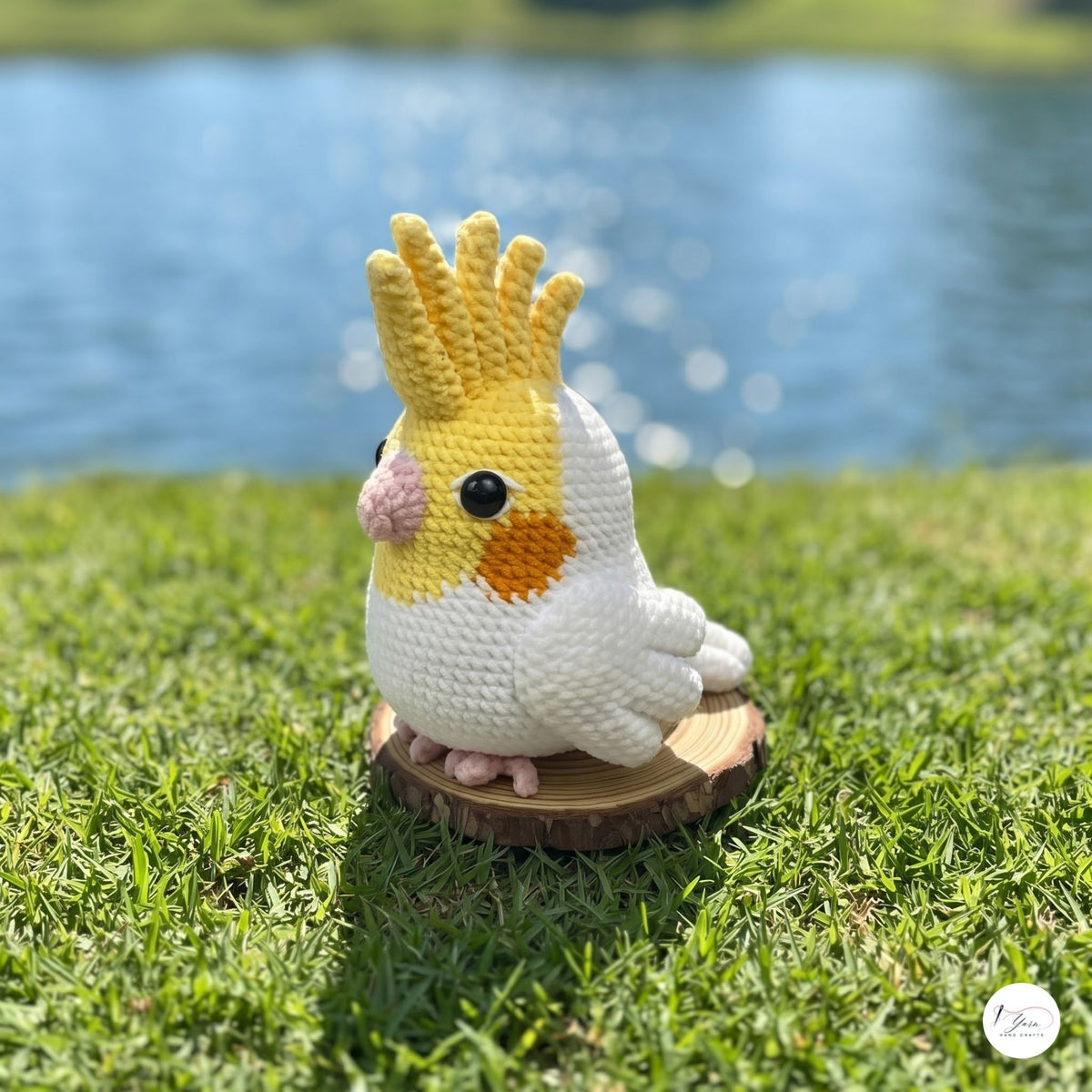 A crocheted amigurumi cockatiel figurine sitting on a small round wooden slice, placed on green grass with a blurred lake and trees in the background.