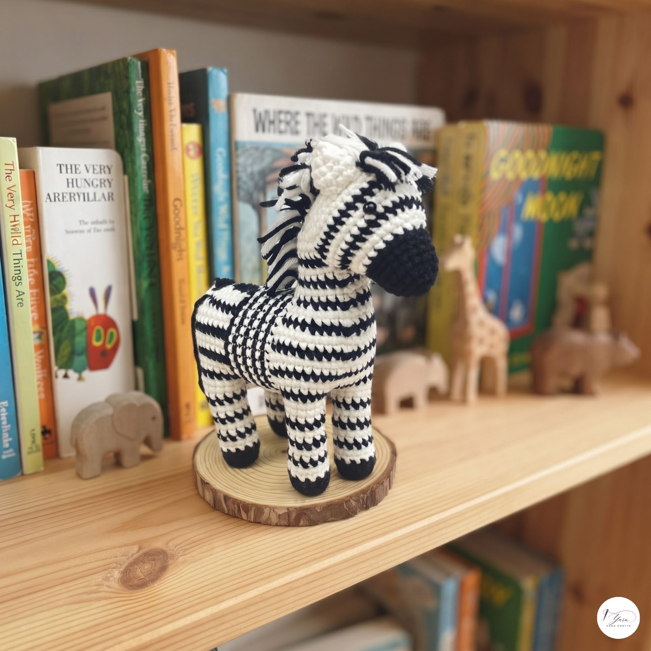 A crocheted zebra toy with black and white striped pattern and a dark blue nose, standing on a round wooden slice base on a wooden bookshelf.