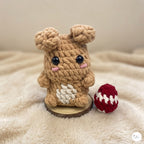 A crocheted plush bunny toy in tan and cream sits on a small wooden base next to a red and white crocheted egg, set against a soft beige blanket background.