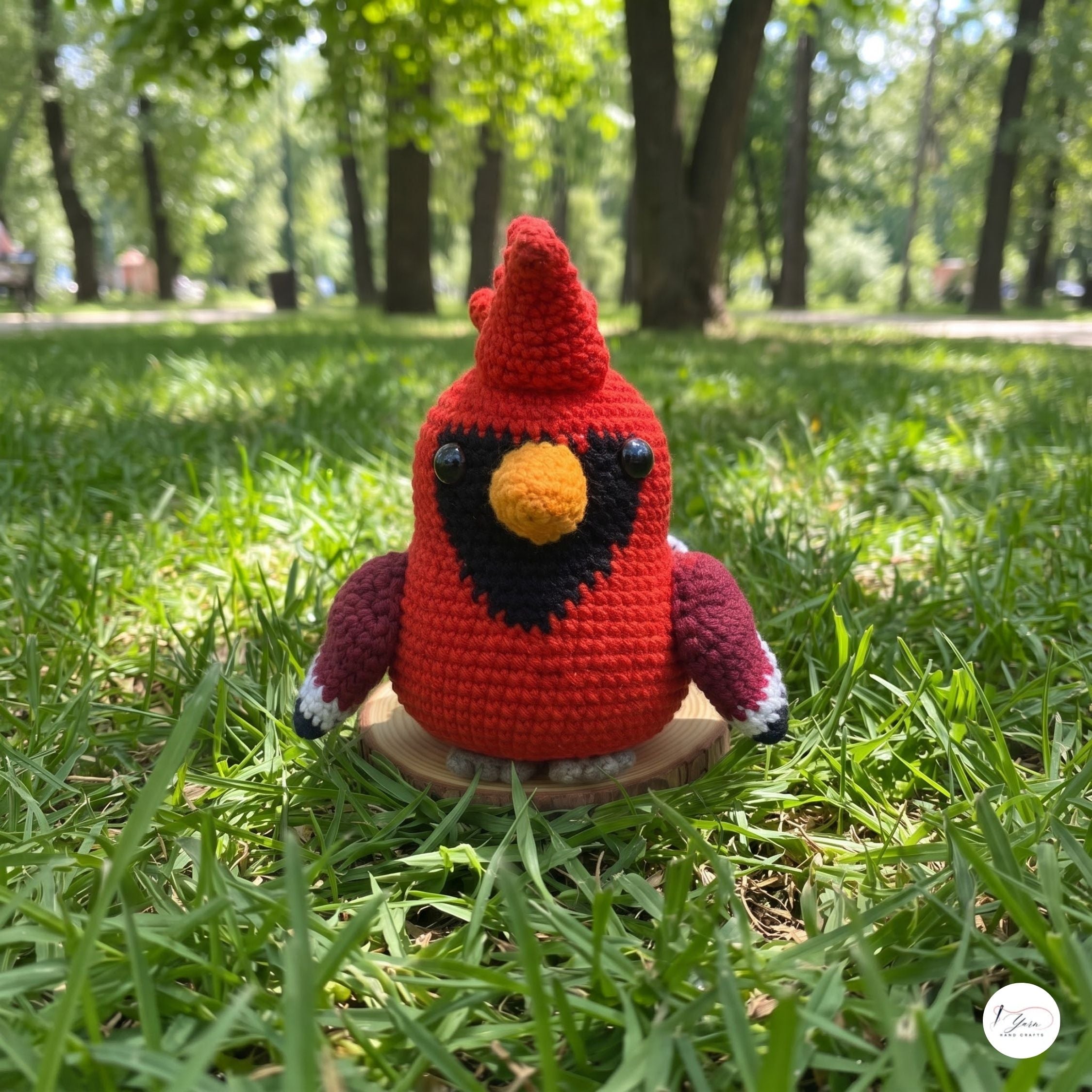 A handmade red crochet cardinal toy sits on a small wooden stand in a grassy park with trees in the background.
