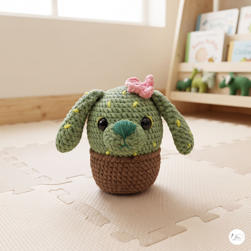 A crocheted plush cactus dog toy with long floppy ears, a pink bow, and a brown pot base, placed on a beige play mat in a softly lit room with a blurred bookshelf in the background.
