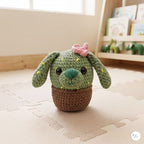 A crocheted plush cactus dog toy with long floppy ears, a pink bow, and a brown pot base, placed on a beige play mat in a softly lit room with a blurred bookshelf in the background.