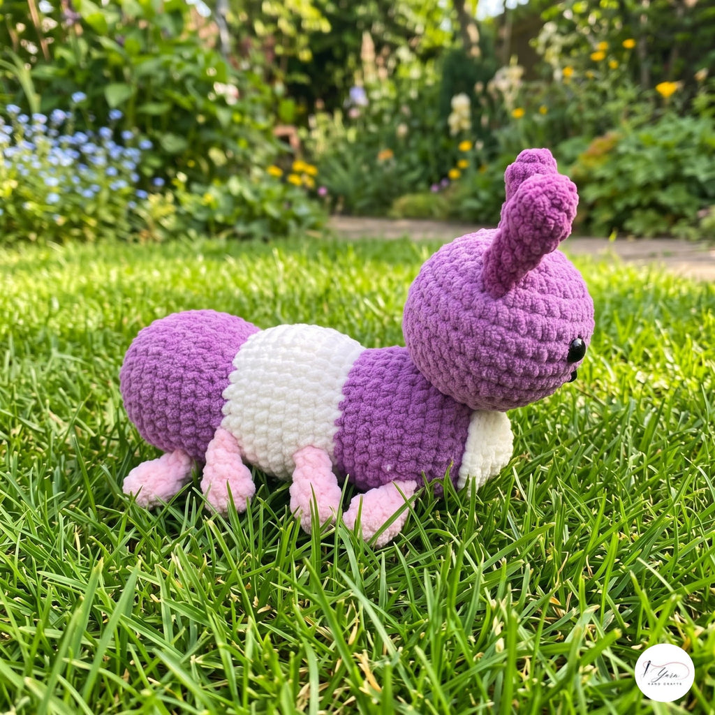 A handmade crocheted caterpillar toy in purple, white, and pink, resting on a green grass lawn with a blurred garden background.