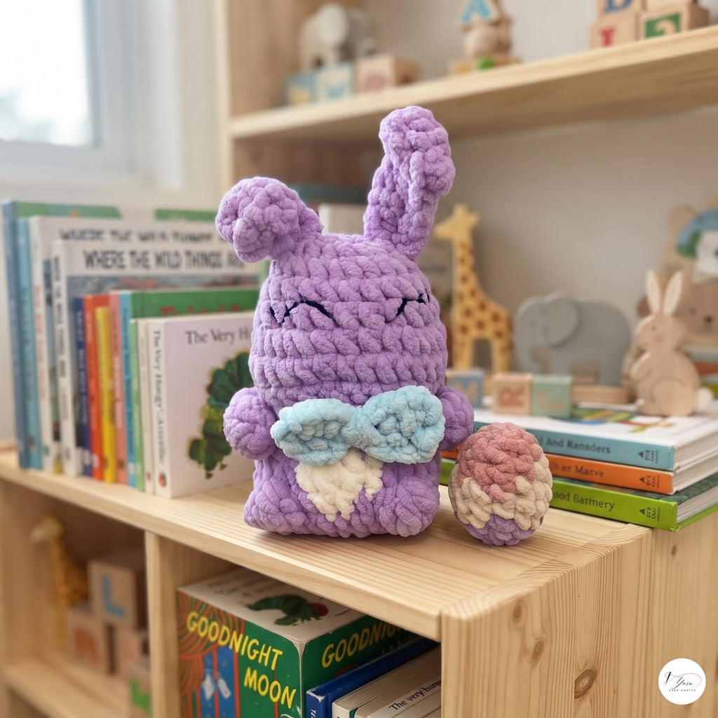 A purple crocheted bunny plush toy with closed eyes and a small light blue and cream bow, sitting on a wooden bookshelf next to a matching crocheted egg. The scene is set in a cozy, brightly lit children's room with books and toys in the background.