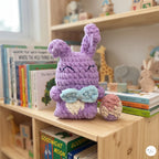 A purple crocheted bunny plush toy with closed eyes and a small light blue and cream bow, sitting on a wooden bookshelf next to a matching crocheted egg. The scene is set in a cozy, brightly lit children's room with books and toys in the background.